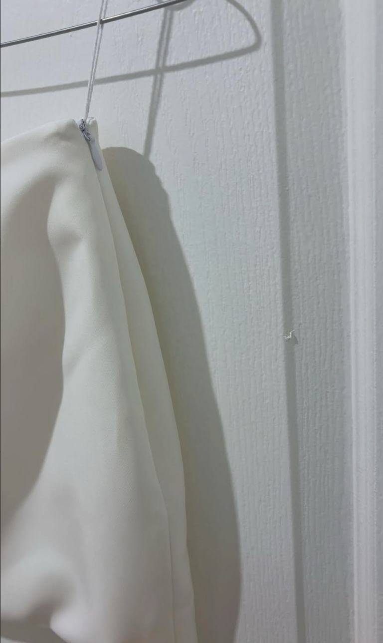 Close up of a zipper on the side of a white mini skirt hanging on a rack against a white wall